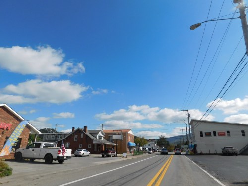 A small town street with shops, parked cars, and a clear blue sky with scattered clouds.
