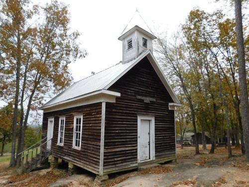 A rustic wooden schoolhouse with a steeple, surrounded by trees and autumn foliage.