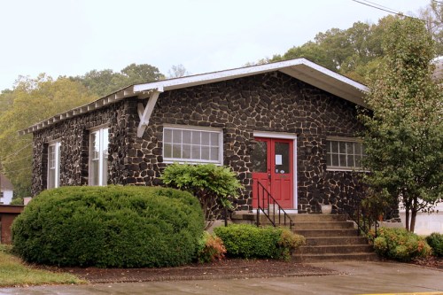 A stone house with a red door, surrounded by greenery and steps leading to the entrance.