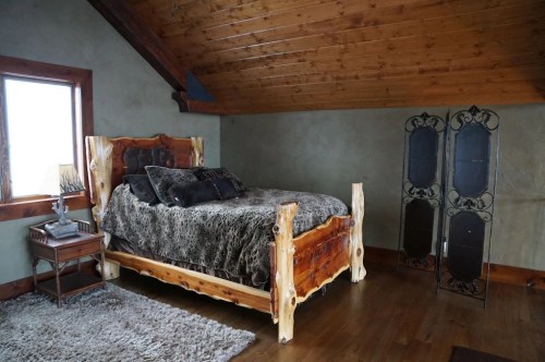 Cozy bedroom featuring a wooden bed, plush bedding, a nightstand, and a decorative screen against a rustic wall.