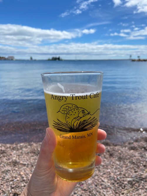 A hand holds a pint glass with "Angry Trout Cafe" logo, overlooking a calm lake and blue sky.