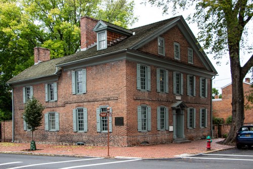 Historic brick building with green shutters, surrounded by trees, located at a street corner.