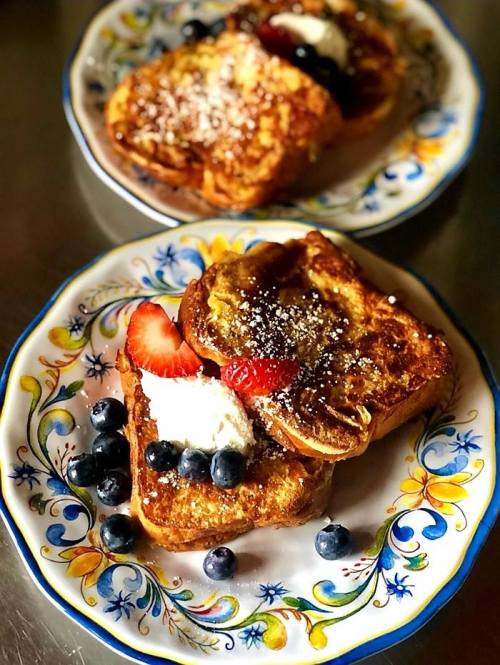 Two plates of French toast topped with strawberries, blueberries, whipped cream, and powdered sugar, on decorative dishes.