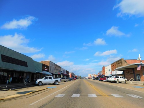 A wide street lined with shops and parked cars under a blue sky with scattered clouds.