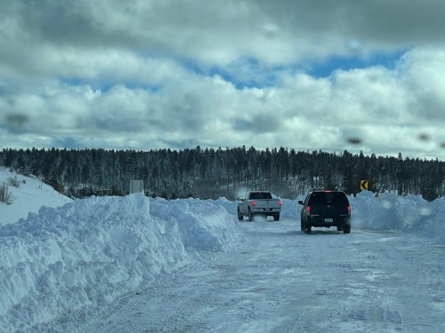 Two vehicles navigate a snowy road surrounded by tall snowbanks and a forest under a cloudy sky.