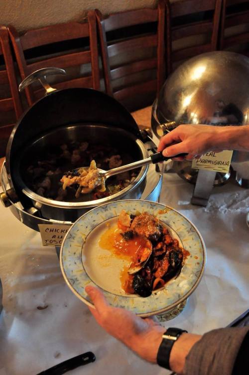 A person serving food from a chafing dish onto a plate, featuring chicken and various side dishes.