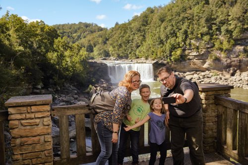A family of four takes a selfie in front of a waterfall, surrounded by lush greenery and a clear blue sky.