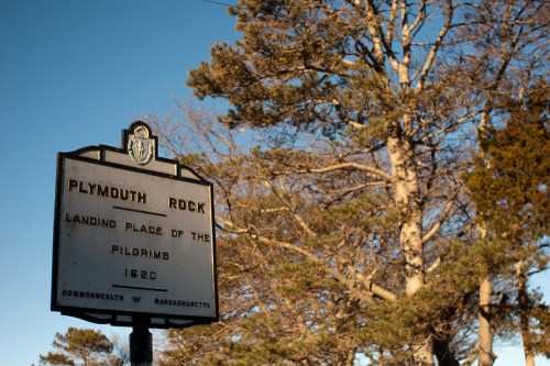 Sign for Plymouth Rock, marking the Pilgrims' landing place in 1620, with trees and clear blue sky in the background.
