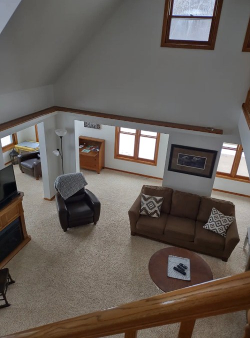 A spacious living room with a high ceiling, featuring a couch, armchair, coffee table, and large windows.