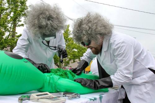 Two scientists in lab coats and wigs examine a green inflatable alien on a table, using medical equipment.