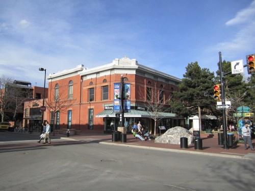 A busy street corner featuring a brick building, trees, and pedestrians near a traffic signal on a clear day.