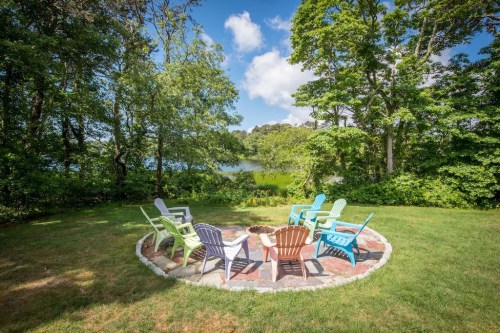 A circular stone patio with colorful chairs surrounds a fire pit, overlooking a serene lake and lush greenery.