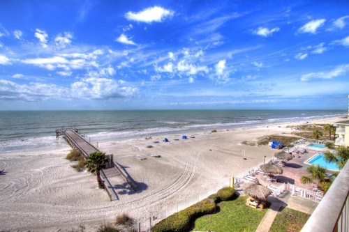 A scenic beach view with a wooden pier, sandy shore, and blue sky dotted with clouds. Sun loungers are visible near the water.
