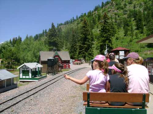 A group of children and adults sit on a train, pointing towards a scenic landscape with trees and model buildings.