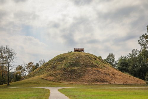 A grassy mound with a wooden observation deck on top, surrounded by trees under a cloudy sky.