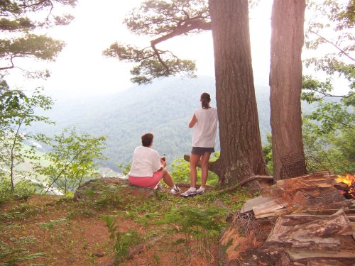 Two people sit on a rocky ledge, overlooking a misty mountain landscape surrounded by trees.