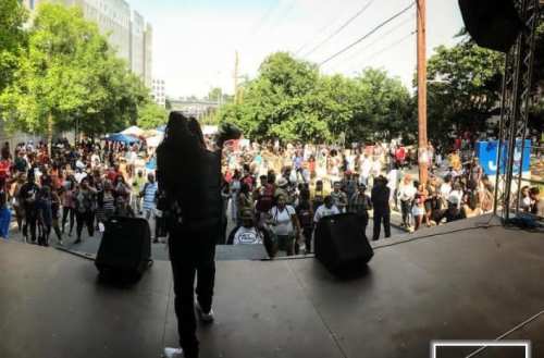 A performer on stage facing a large crowd at an outdoor event, surrounded by trees and buildings.
