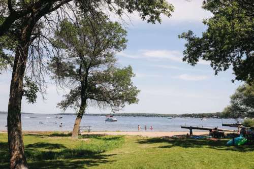 A serene lakeside view with trees, a sandy beach, and people enjoying water activities under a clear blue sky.