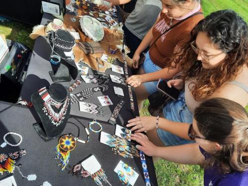 Three people examine colorful handmade jewelry and crafts displayed on a table at an outdoor market.