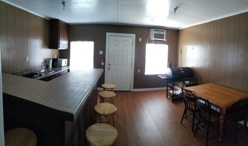 A spacious kitchen with a bar, stools, dining table, and an air conditioner, featuring brown walls and wooden flooring.
