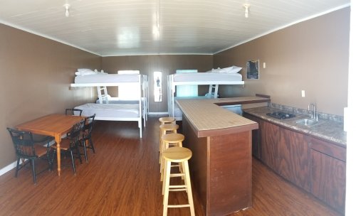Interior of a room with two bunk beds, a dining table, and a kitchenette with bar stools. Warm brown walls and wooden flooring.