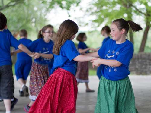 Children in blue shirts and colorful skirts dance together outdoors, smiling and enjoying a lively group activity.