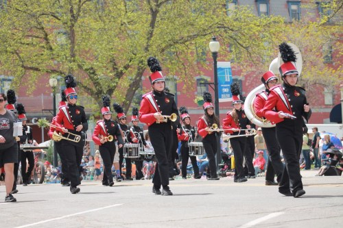 A marching band in red uniforms parades down a street, playing instruments with spectators in the background.