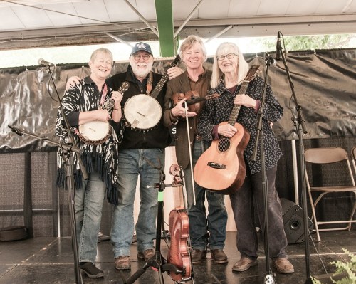 Four musicians stand together on stage, each holding an instrument, smiling at the camera.