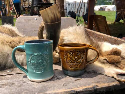 Two ceramic mugs with "Appalachian Festival" designs sit on a wooden table, surrounded by fur and rustic decor.
