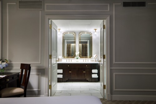 A luxurious hotel bathroom with double sinks, elegant mirrors, and soft lighting, viewed from a bedroom doorway.