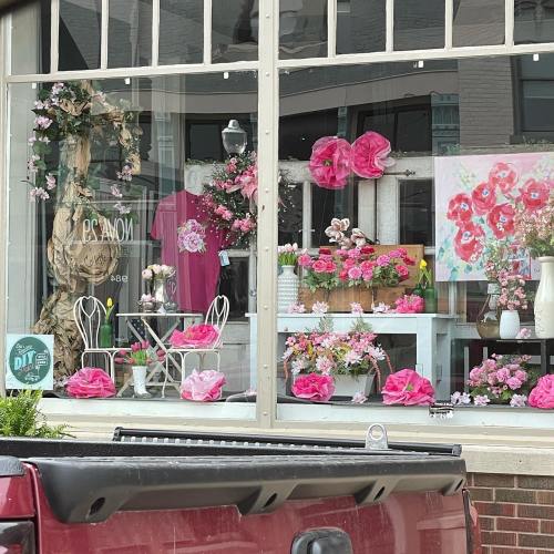 A storefront display featuring pink flowers, decorative items, and a charming seating area.