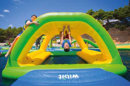 Two children play on a colorful inflatable water structure, one hanging upside down while the other climbs.