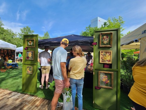 A couple views artwork at an outdoor market, surrounded by tents and greenery under a clear blue sky.