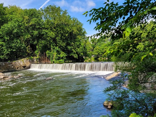 A serene river scene featuring a waterfall surrounded by lush green trees under a clear blue sky.