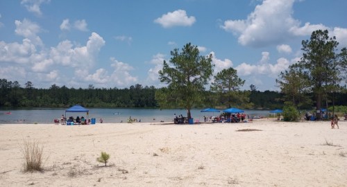 A sandy beach with people enjoying the water, shaded by blue tents under a bright sky with fluffy clouds.