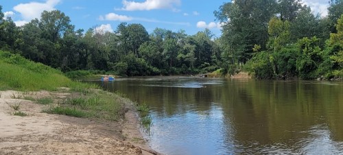 A serene river scene with lush greenery, a sandy bank, and a clear blue sky with fluffy clouds.