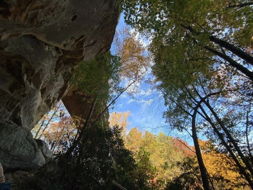 View from below a rocky overhang, framed by colorful autumn trees and a clear blue sky.