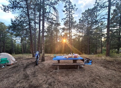 A campsite with a picnic table covered in food, surrounded by trees, as the sun sets in the background.