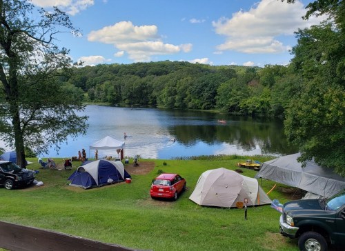 A serene lakeside campsite with tents, cars, and people enjoying the outdoors under a blue sky with fluffy clouds.