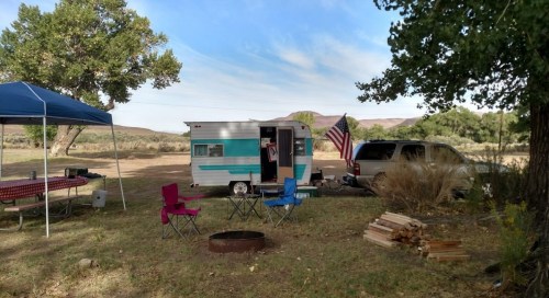 A campsite with a vintage camper, chairs, a fire pit, and an American flag, surrounded by trees and mountains.