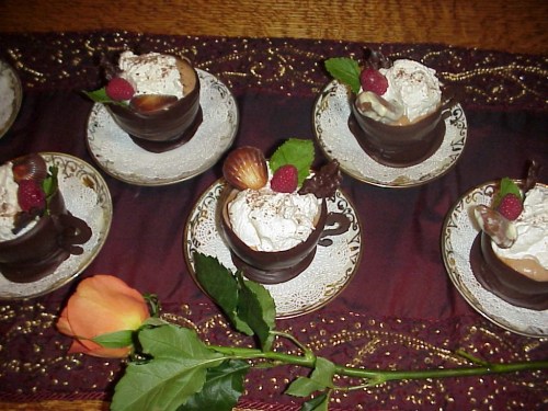 Elegant dessert cups made of chocolate, filled with cream and topped with raspberries, mint, and a rose beside them.