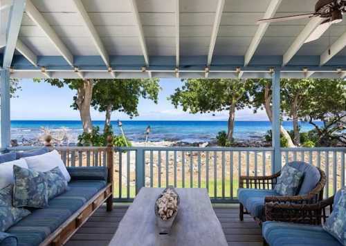 Cozy porch with blue furniture overlooking a serene beach and ocean, framed by lush green trees.