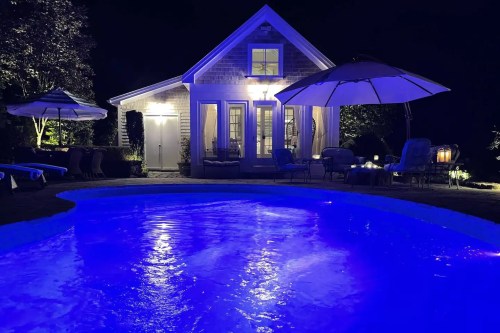 A serene night scene featuring a glowing blue pool and a cozy house with an umbrella and outdoor seating.