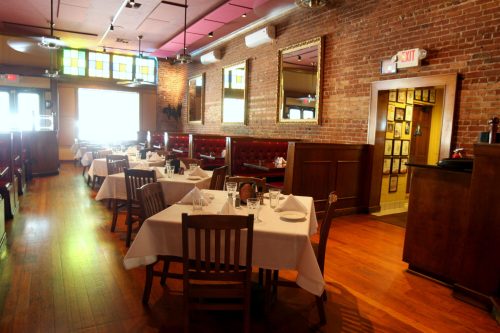 Cozy restaurant interior with wooden tables, white tablecloths, and brick walls, featuring mirrors and colorful windows.