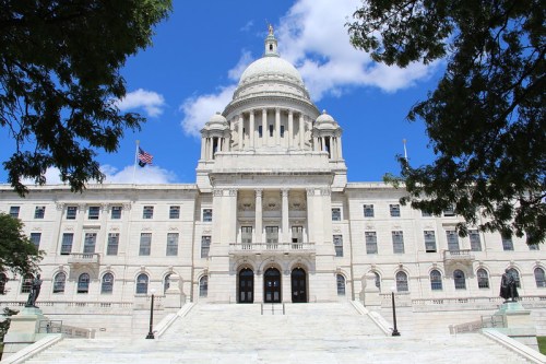The Rhode Island State House, featuring a large dome and steps, set against a blue sky with scattered clouds.
