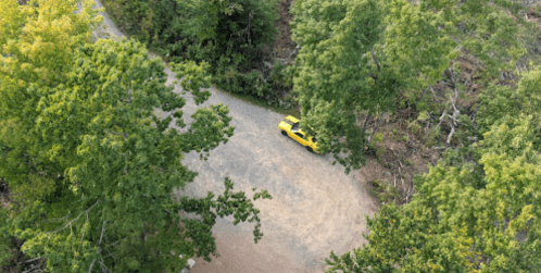 Aerial view of a yellow car parked on a gravel road surrounded by green trees.