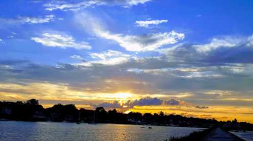 A serene sunset over a calm waterway, with colorful clouds and silhouettes of trees and boats in the foreground.