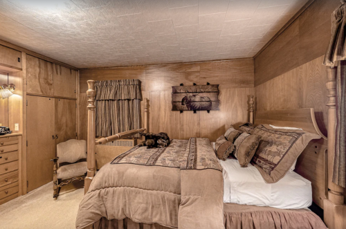 Cozy bedroom with wooden walls, a large bed with patterned bedding, and a chair in the corner. A dog rests on the bed.