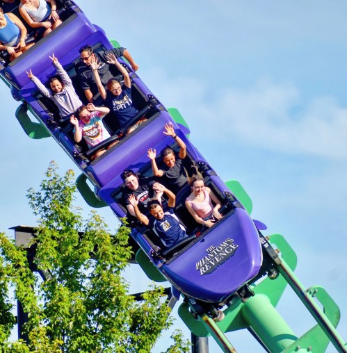 A roller coaster with riders raising their hands in excitement as it climbs a steep incline against a blue sky.