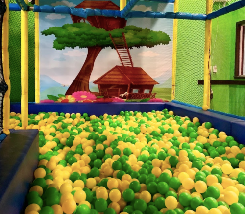 A colorful ball pit filled with green, yellow, and blue balls, set against a playful treehouse backdrop.